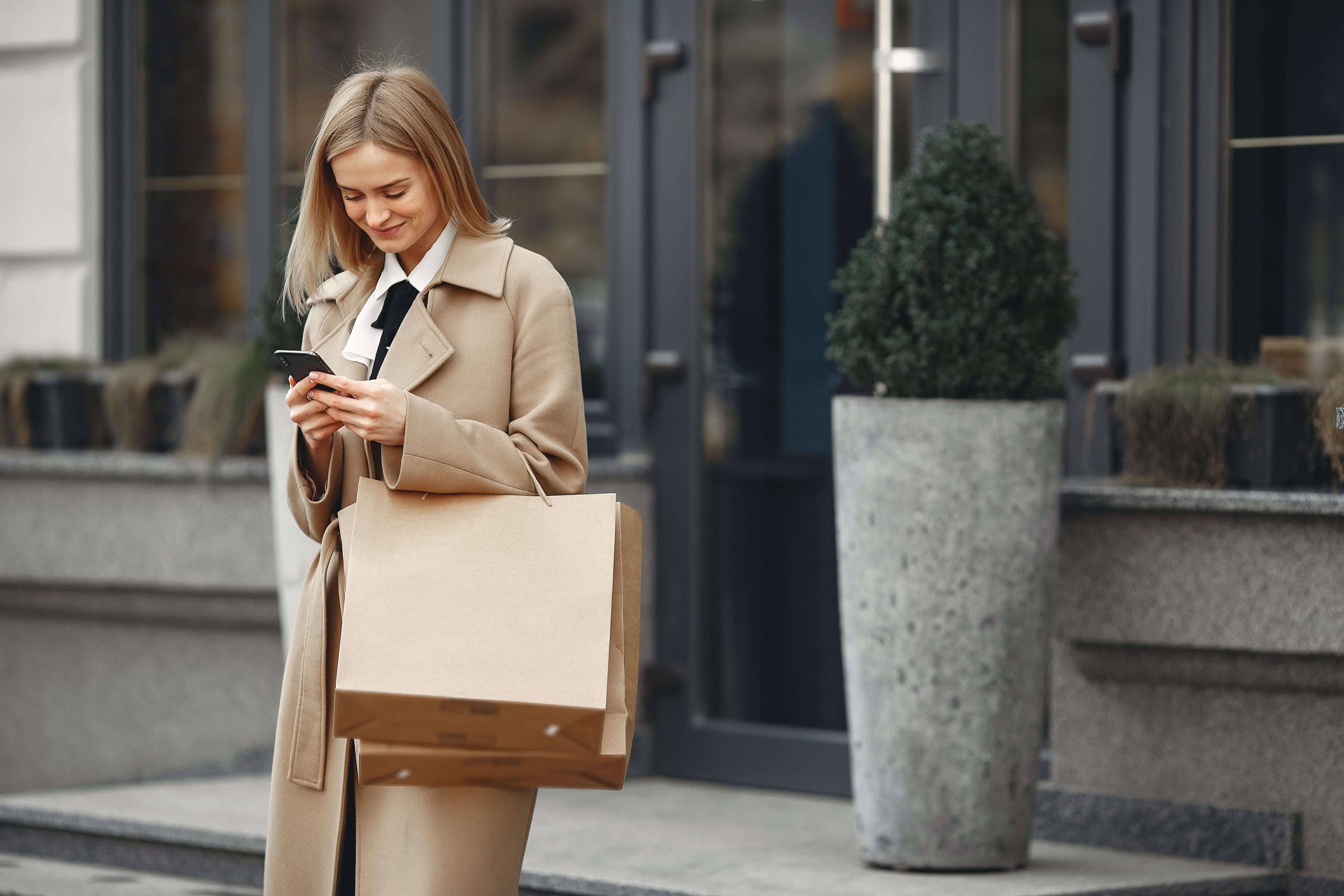 woman with paper bags standing on sidewalk and typing on cell phone